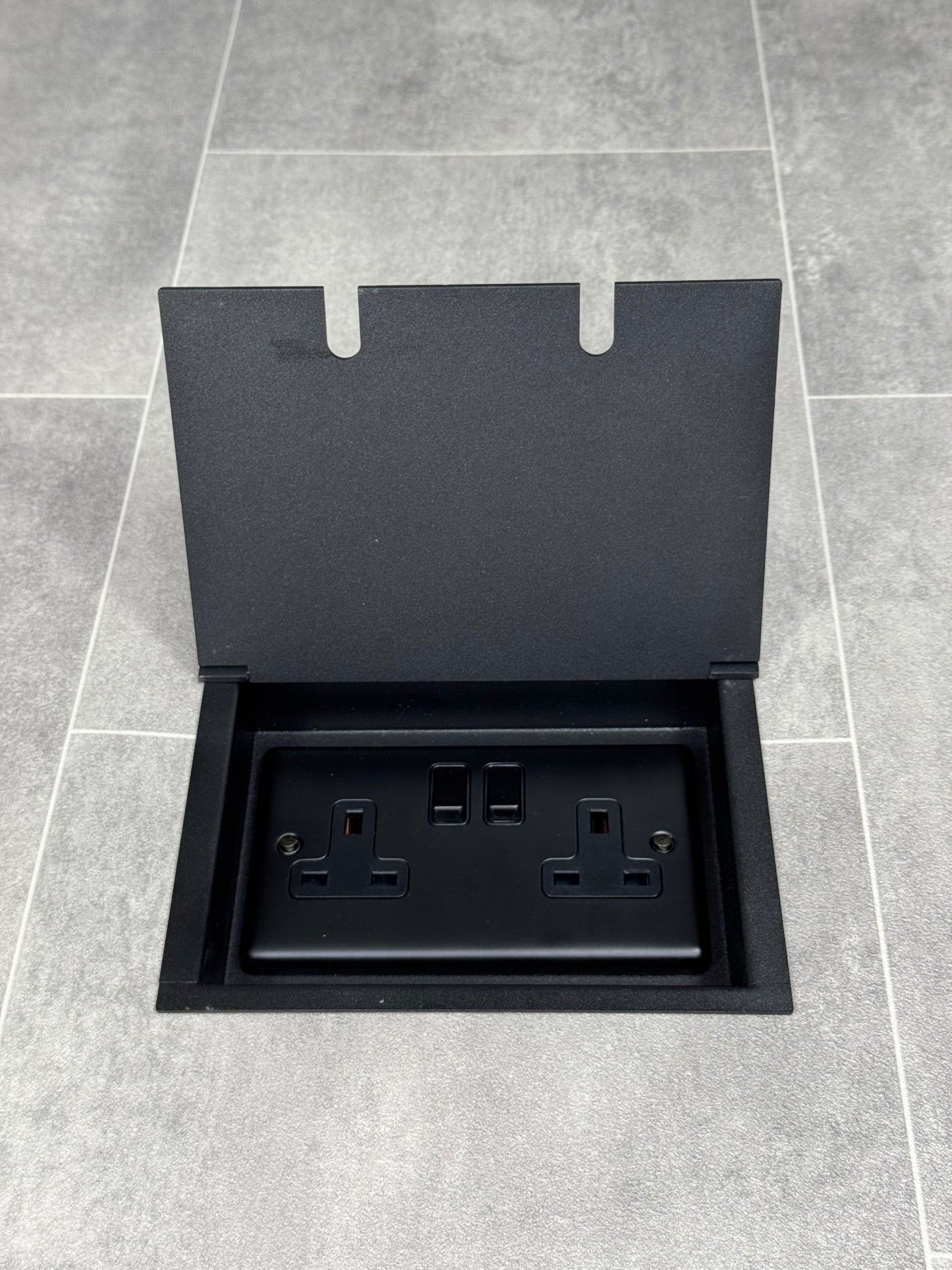A matt black double recessed floor socket with a 13amp switched plug socket, in a tiled floor.
