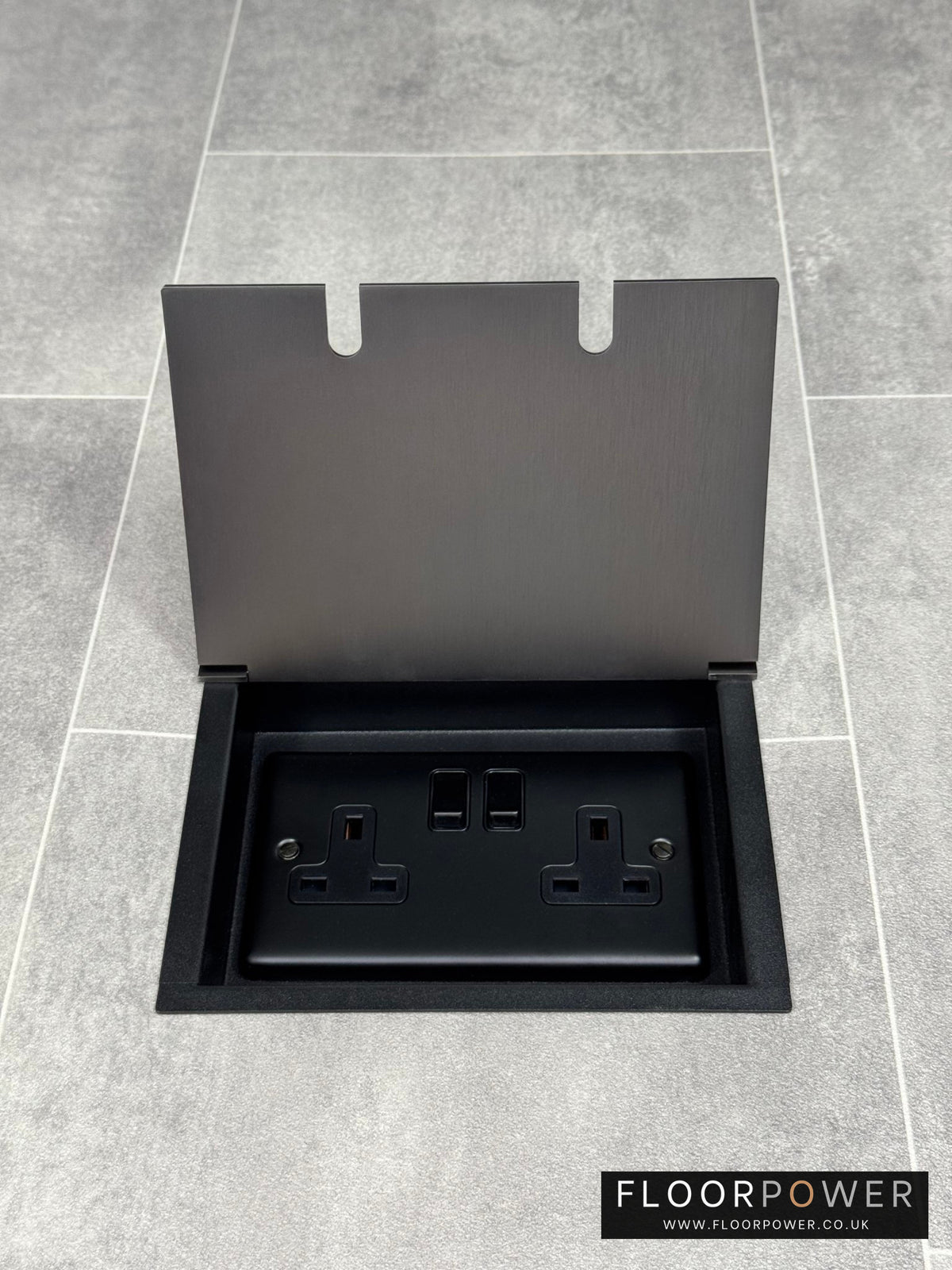 Am old bronze double recessed floor socket, with its cover open, revealing a double 13 amp switched plug socket.