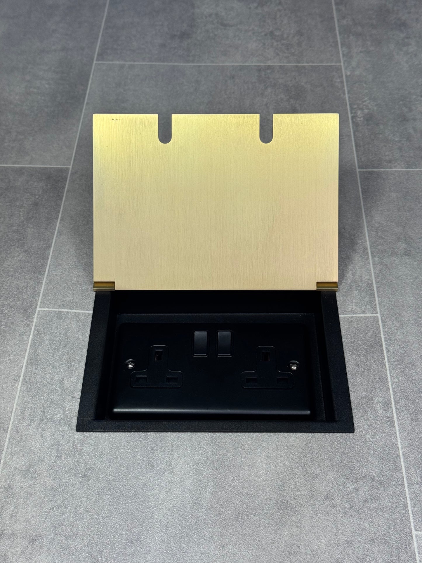 A satin brass double recessed floor socket with twin 13 amp switched sockets in a black base, installed in a floor.