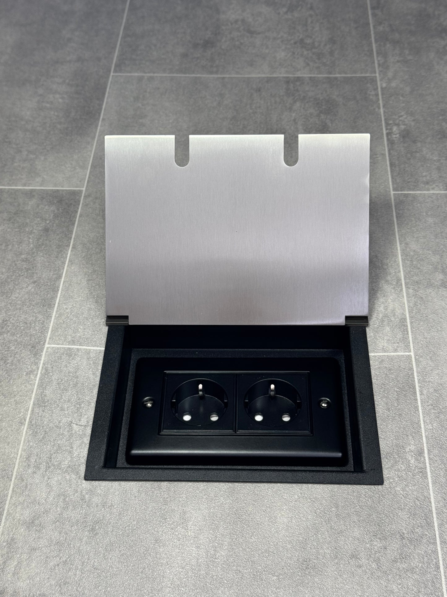 A satin stainless steel double recessed floor socket with two 13amp unswitched Schuko plug sockets, on a tiled floor.