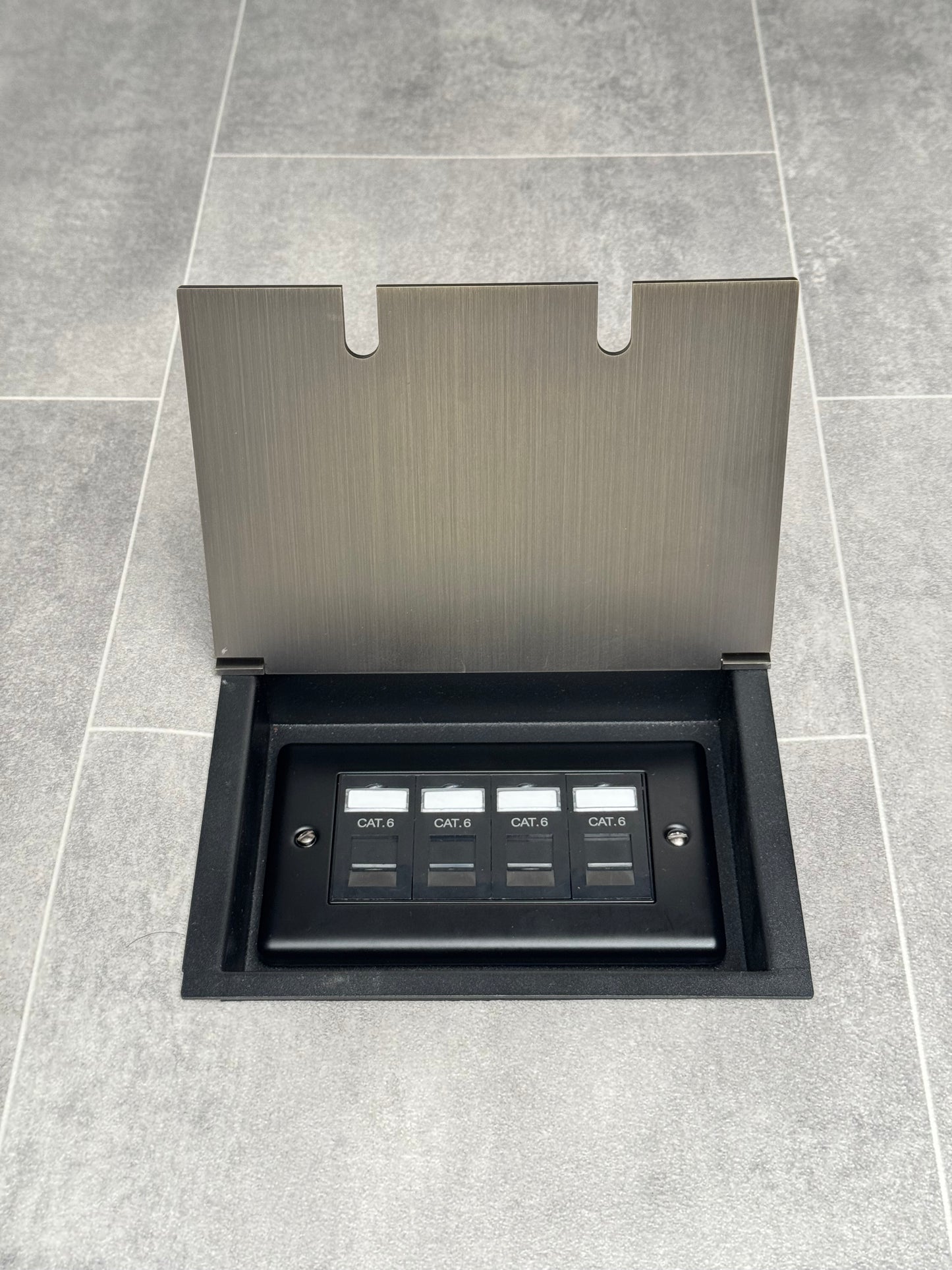 A recessed floor socket in antique brass with a double box design, including 4 gang RJ45 cat6 sockets, opened and set in a tiled floor.