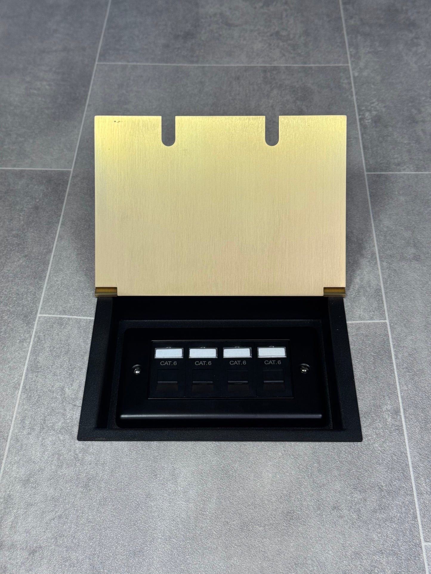 A satin brass double recessed floor socket with a black base installed on a grey floor, opened to show the 4 gang RJ45 cat6 socket.