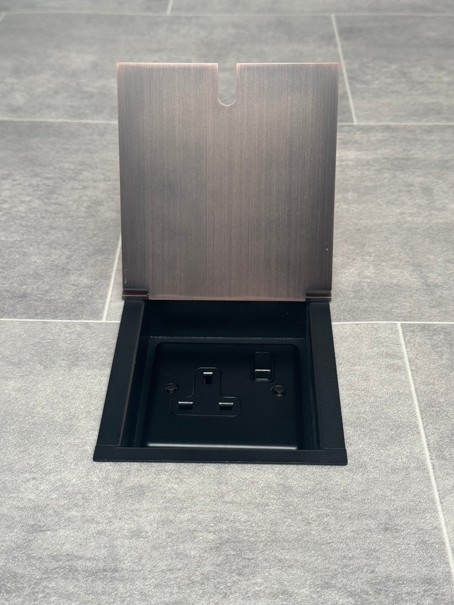 A recessed floor socket with a antique copper finish, opened to show the 13 Amp plug socket.