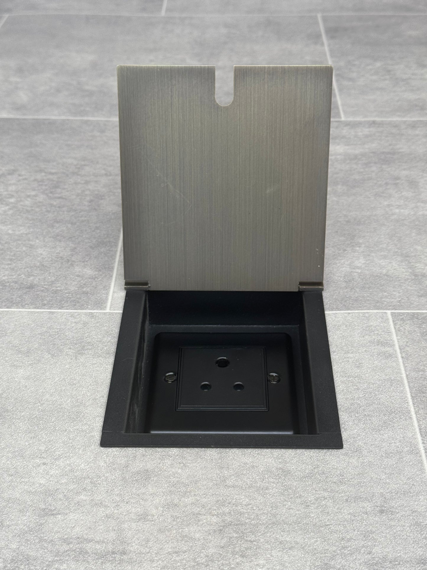 A 5 amp round pin recessed floor socket for lighting with antique brass finish cover, set against a grey tiled floor.