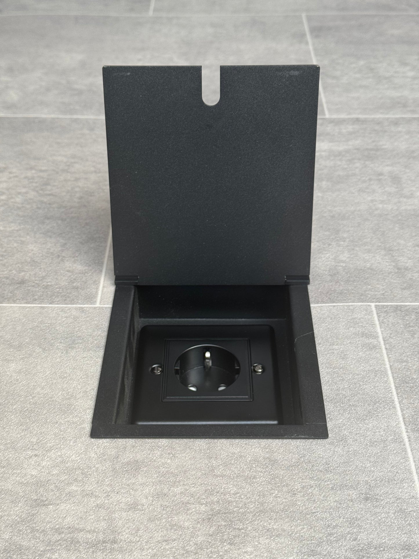 A matt black recessed floor socket with a 13amp Schuko Socket, shown in an open position in a grey floor tiles.
