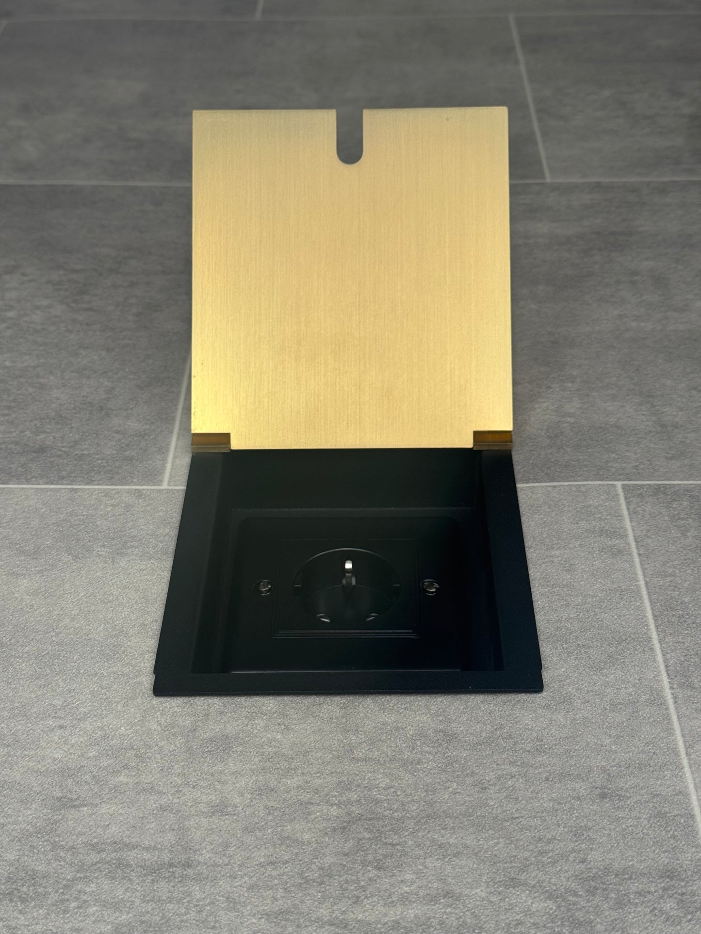 A satin brass single recessed floor socket with a Schuko (European) socket in a black base installed in a floor.