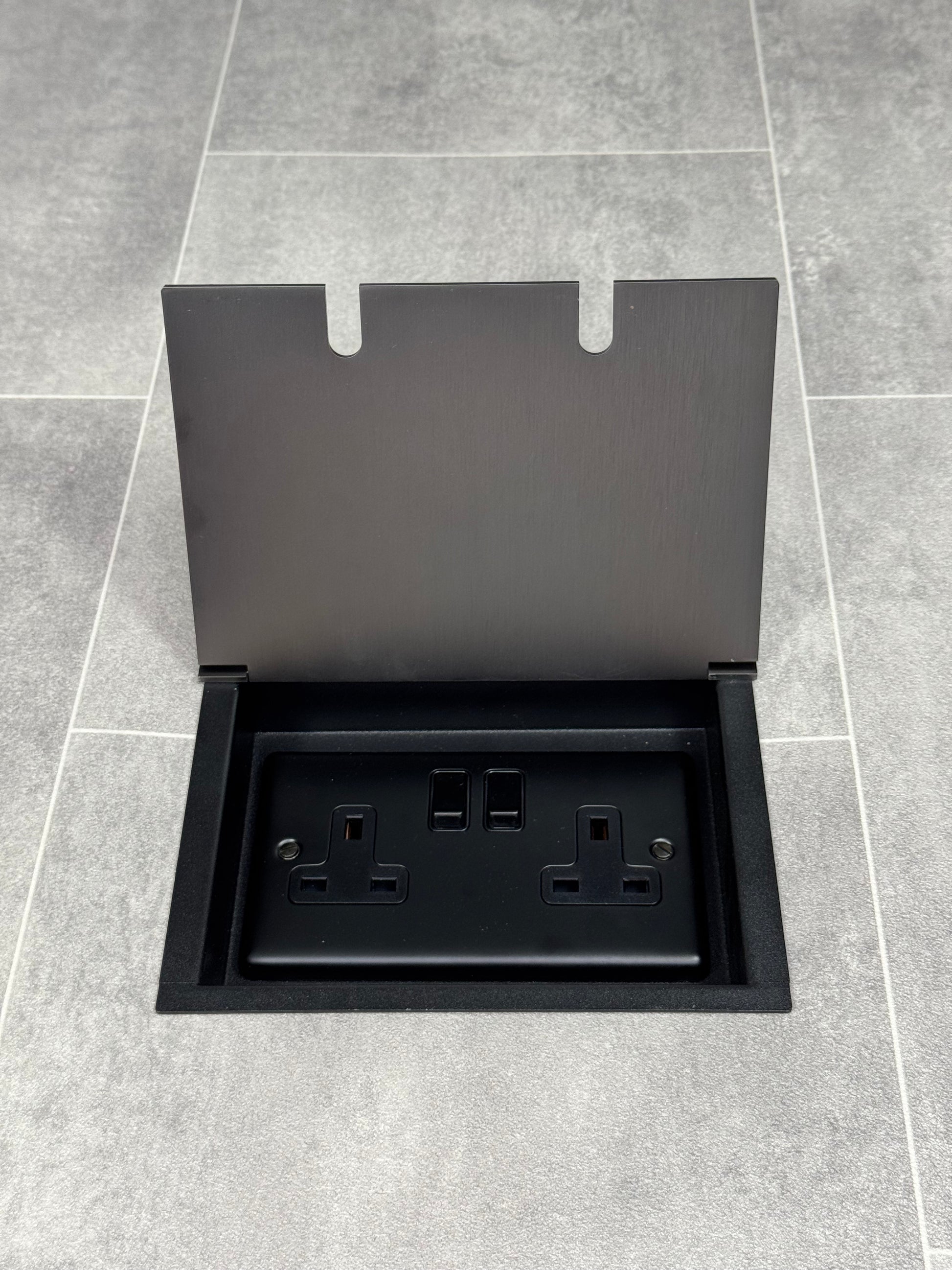 Am old bronze double recessed floor socket, with its cover open, revealing a double 13 amp switched plug socket.