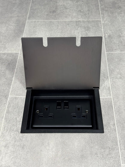 Am old bronze double recessed floor socket, with its cover open, revealing a double 13 amp switched plug socket.