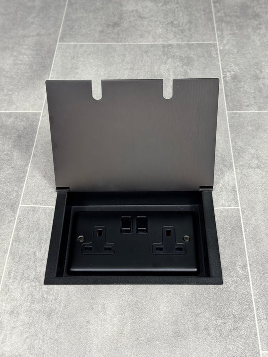 Am old bronze double recessed floor socket, with its cover open, revealing a double 13 amp switched plug socket.