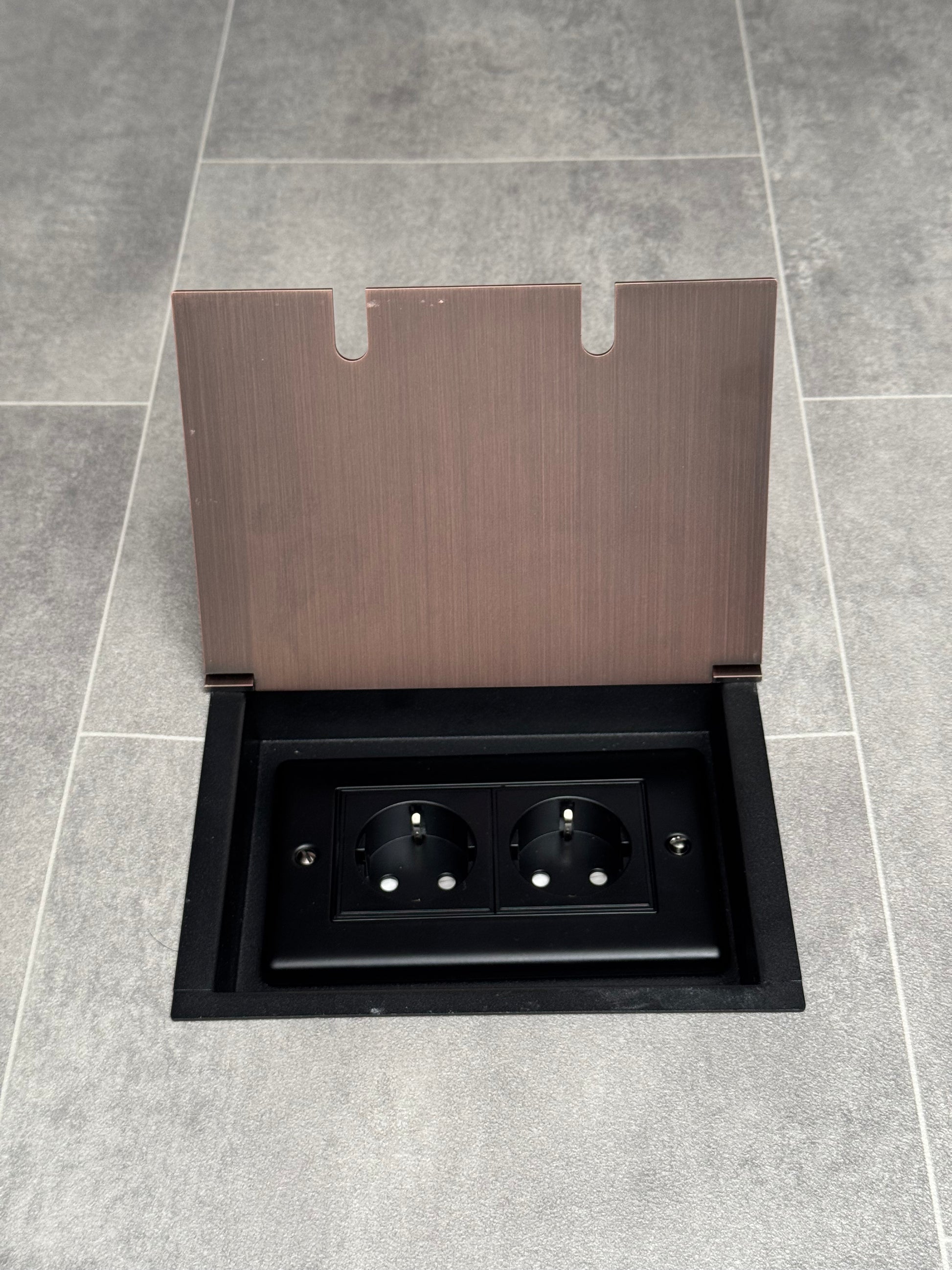 Antique copper double recessed floor socket with two Schuko (European) sockets in a black base installed in a floor.