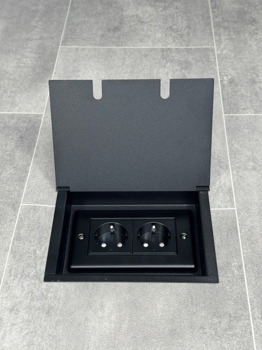 A matt black recessed floor socket  opened to reveal a double 13 amp unswitched Schuko socket.