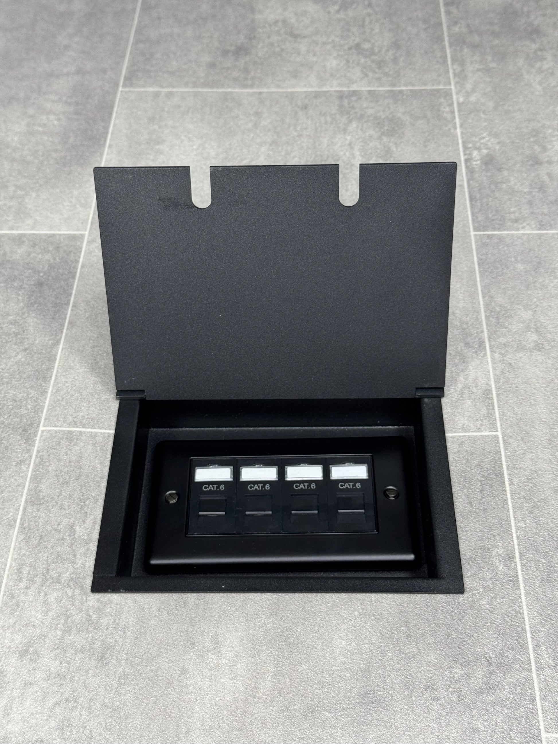 A matt black double recessed floor socket with a black base installed on a grey floor, opened to show the 4 gang RJ45 cat6 socket.