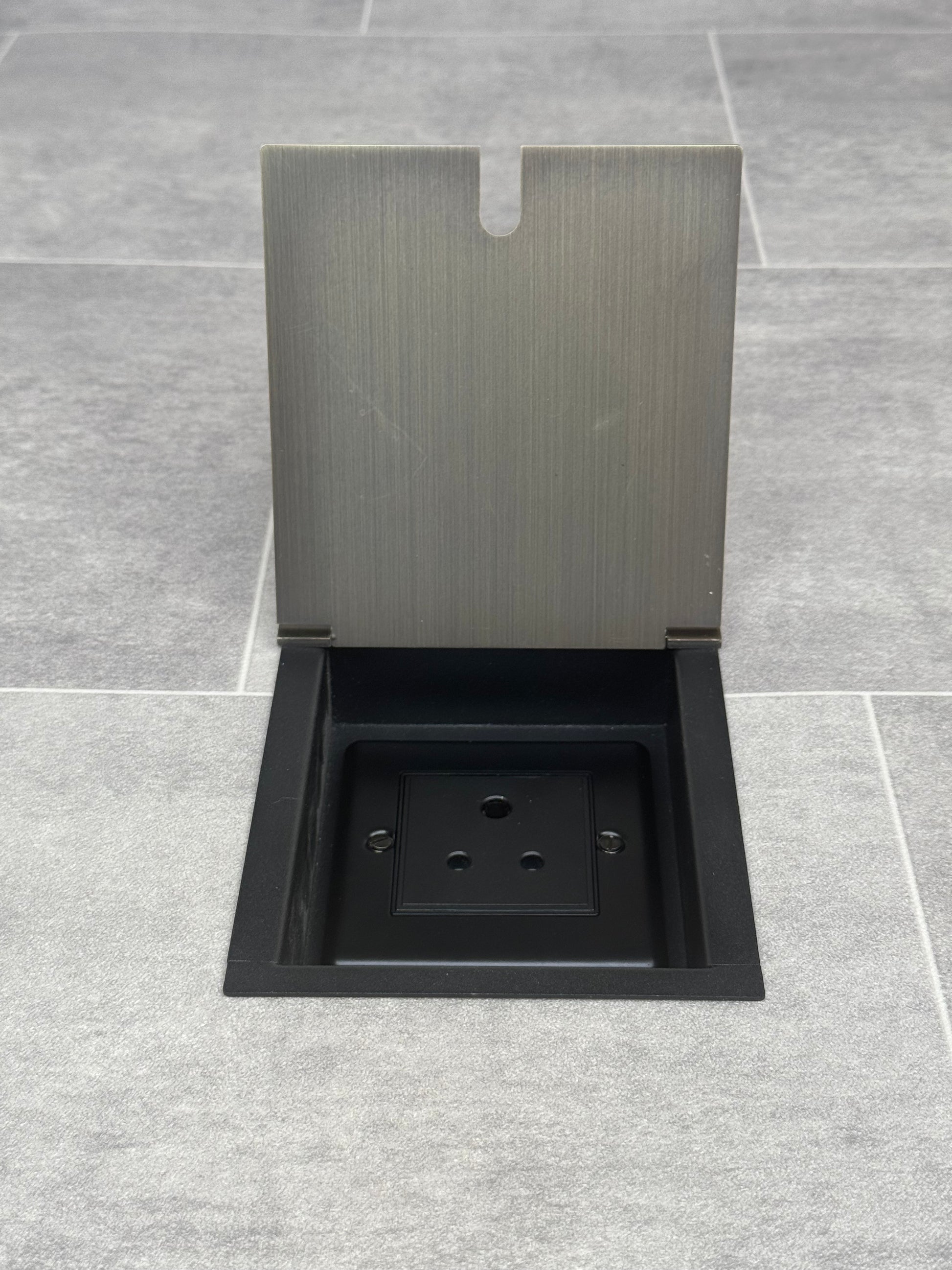 A 5 amp round pin recessed floor socket for lighting with antique brass finish cover, set against a grey tiled floor.