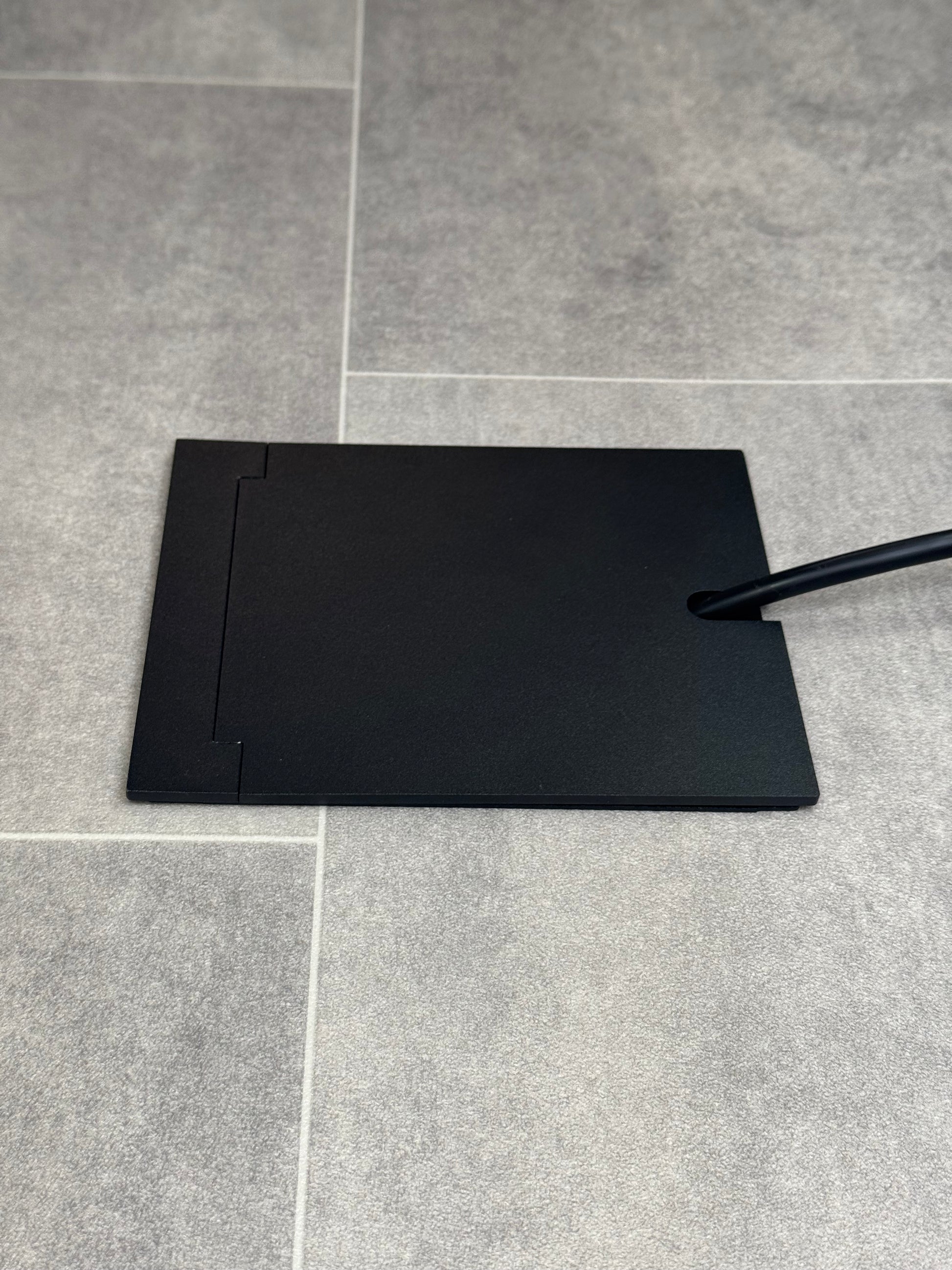 Single Matt Black Recessed Floor Socket With Lid Closed Flat Showing Just Cable Coming Out If The Socket - Side On View.