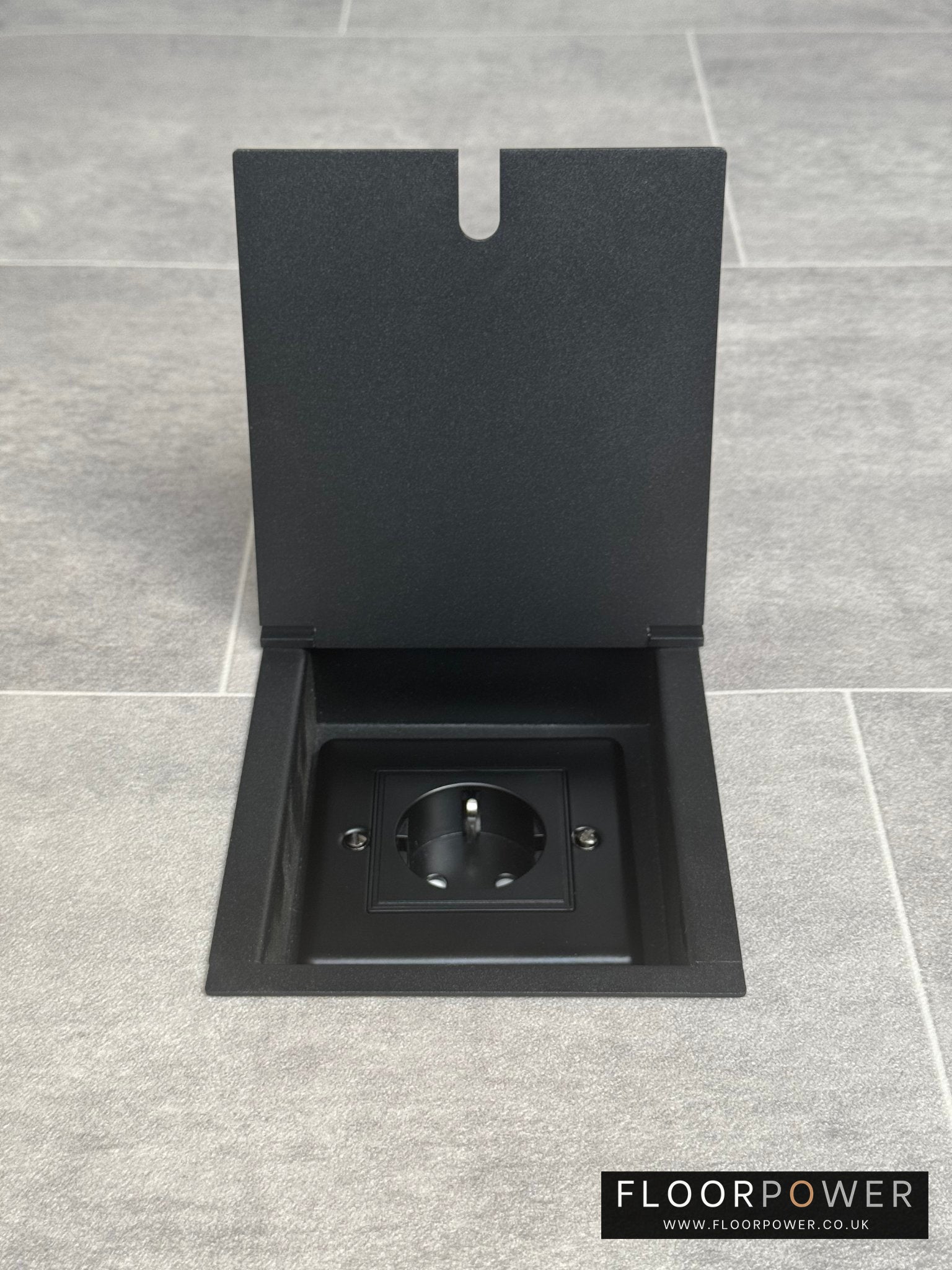 A matt black recessed floor socket with a 13amp Schuko Socket, shown in an open position in a grey floor tiles.