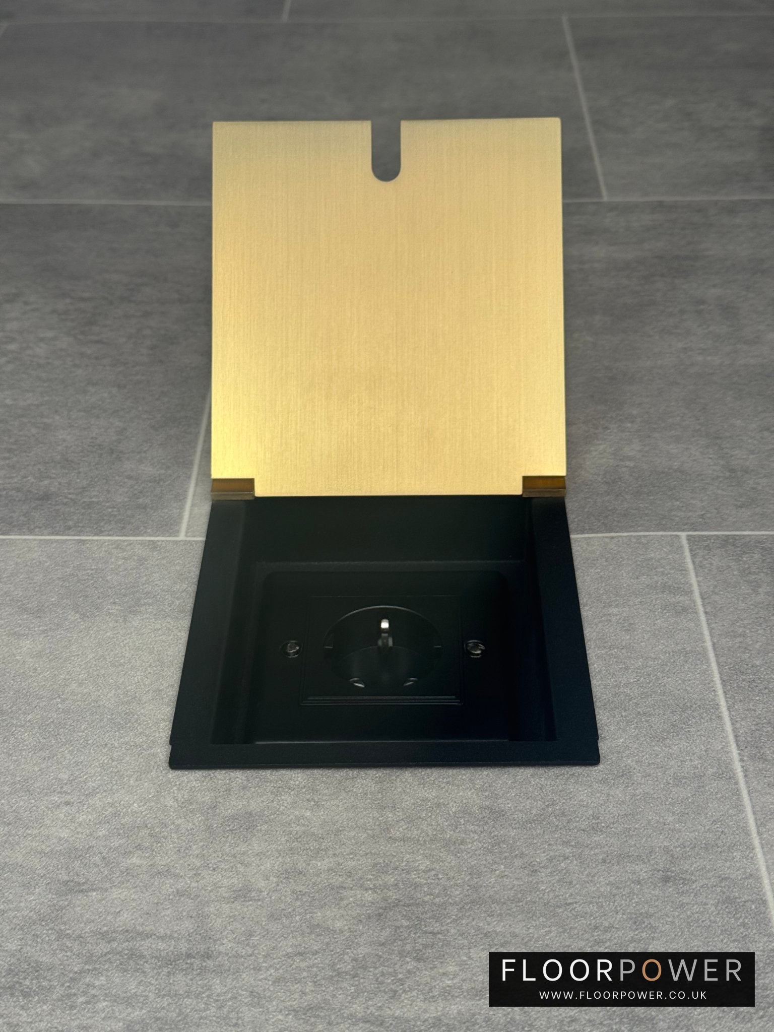 A satin brass single recessed floor socket with a Schuko (European) socket in a black base installed in a floor.