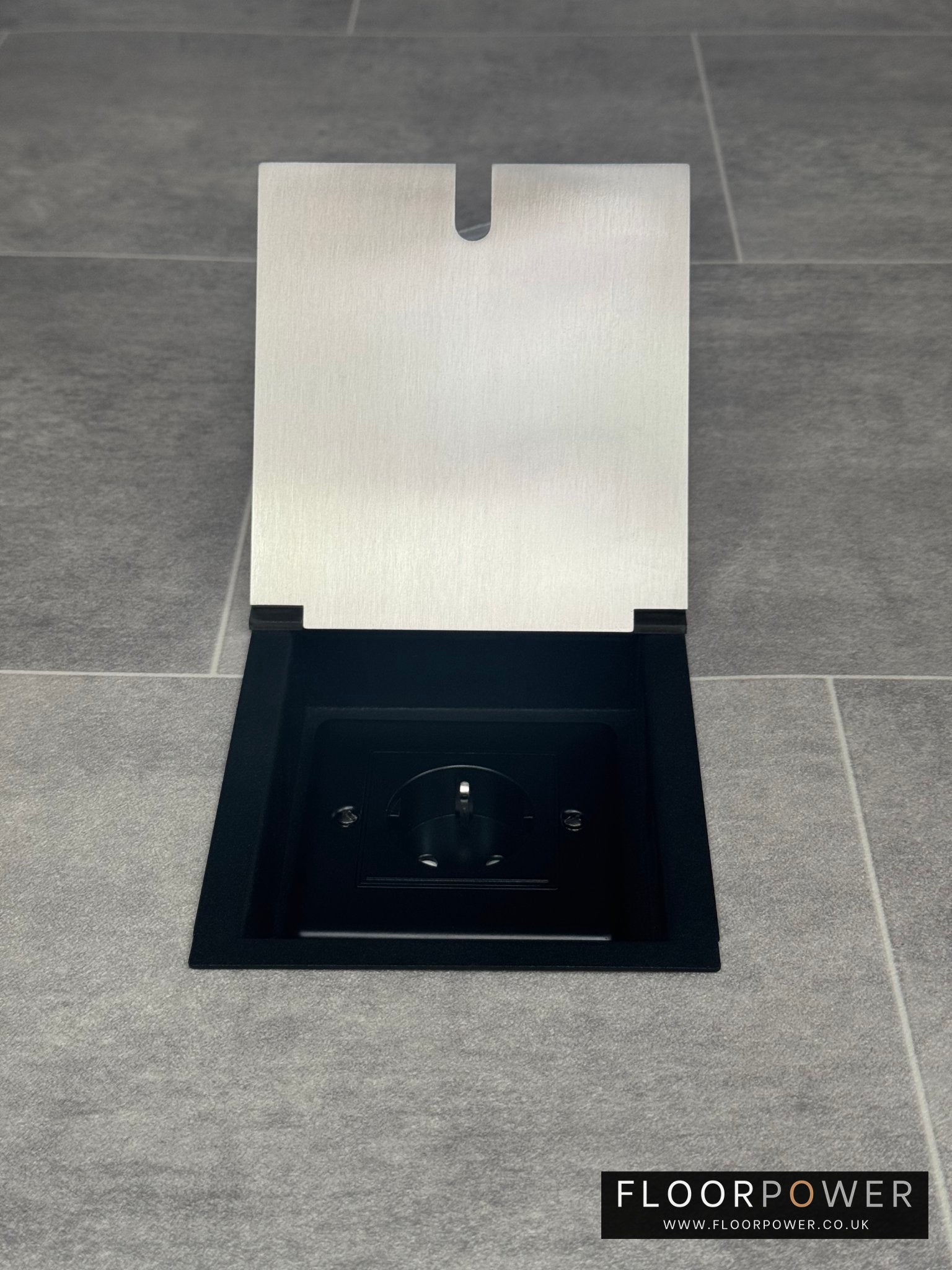 A single satin stainless steel recessed floor socket with a a Schuko (European) plug socket, in a grey tiled floor.