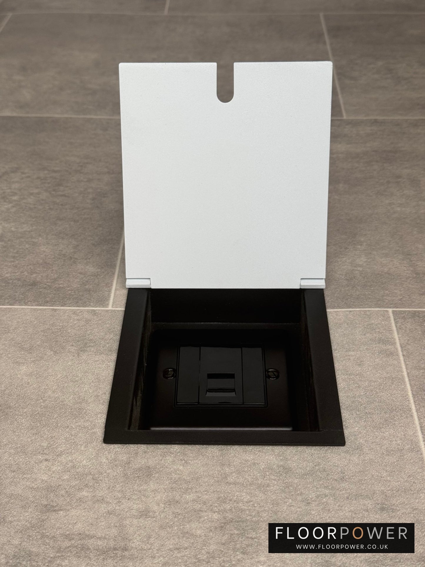A white recessed floor socket with a UK phone socket installed on a tiled floor.